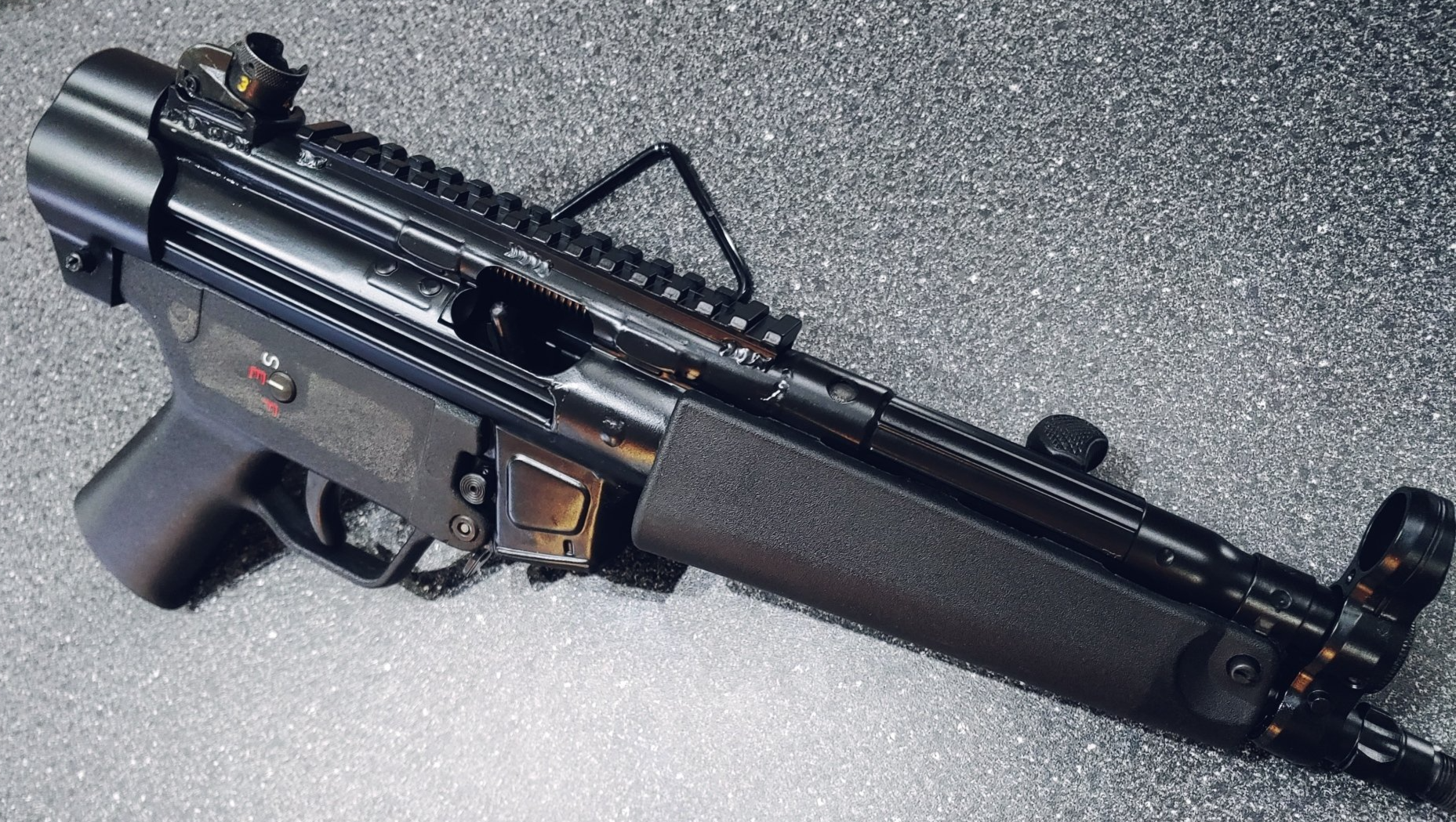 Black submachine gun with a textured handguard, metal rails, and iron sights on a reflective surface.