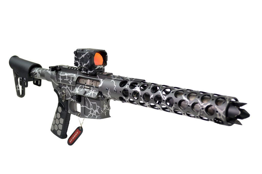 A camo-patterned firearm with a red dot sight and a perforated barrel.