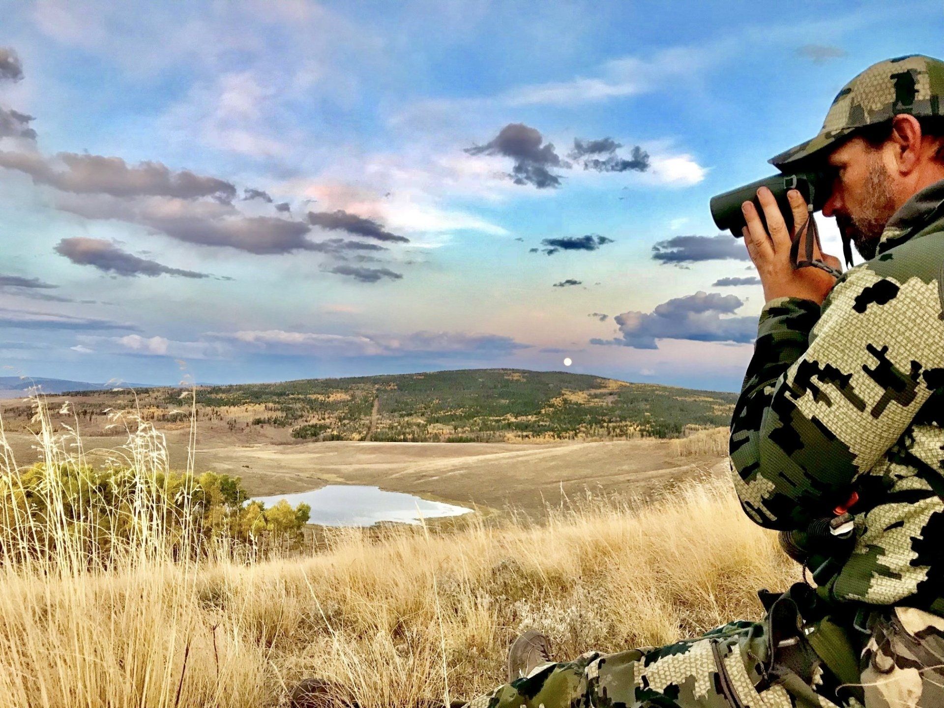 Hunter in camouflage uses binoculars, overlooking a landscape with a pond and mountains under a cloudy sky.