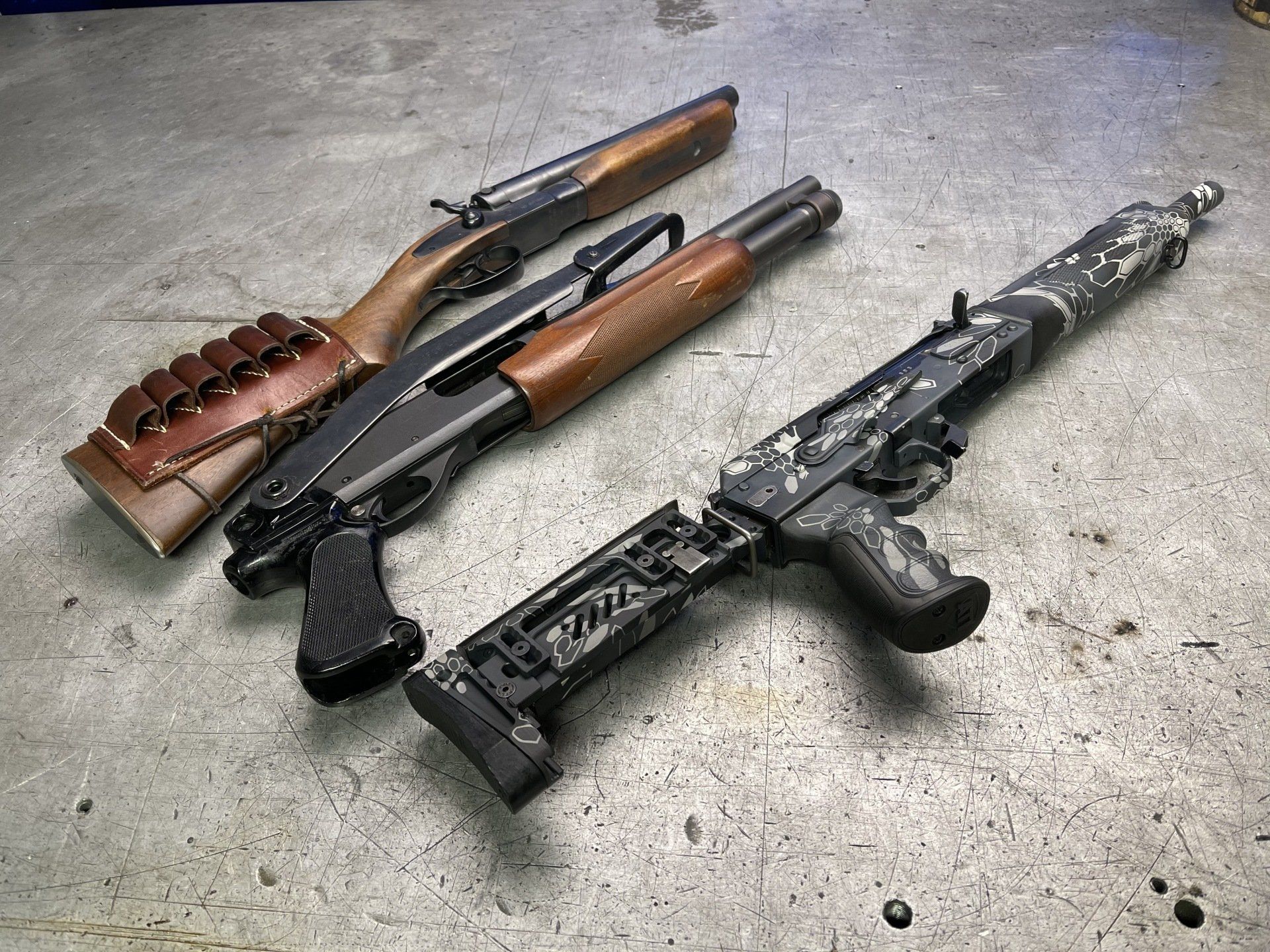 Three shotguns on a metallic surface. Two have wooden furniture; one has camo finish.