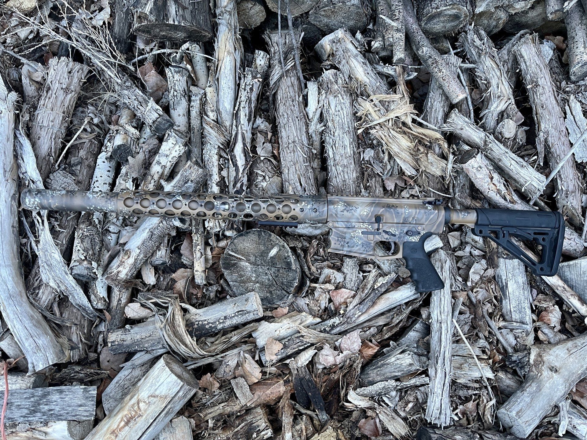 A camouflage patterned firearm resting on a pile of weathered wood.