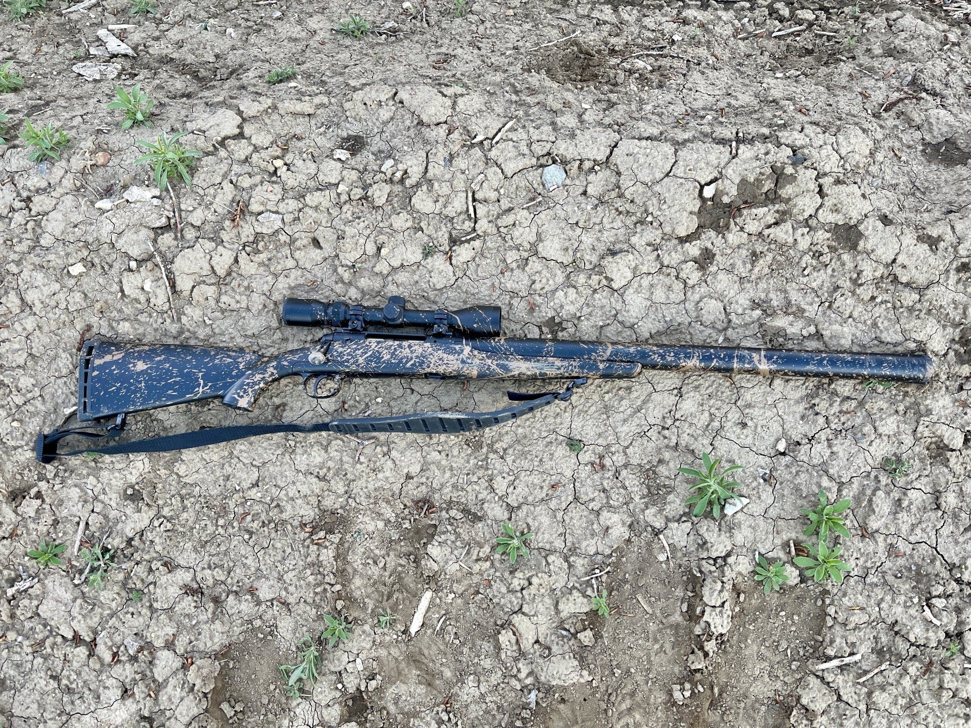 Black rifle with scope and strap on cracked earth.