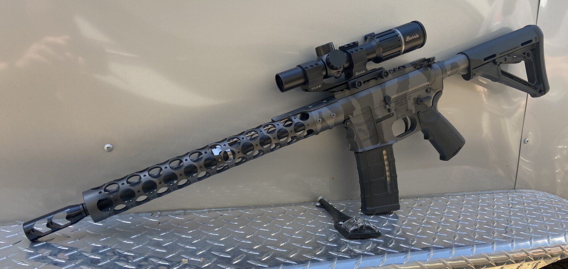 An AR-15 style rifle with a scope. It is resting on a metallic surface with the bolt open.