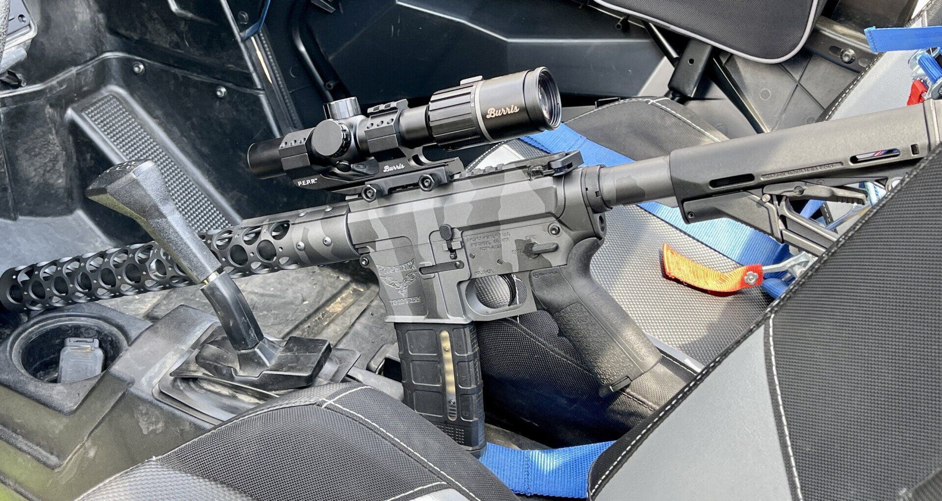 Black rifle with scope and magazine rests in a vehicle interior.