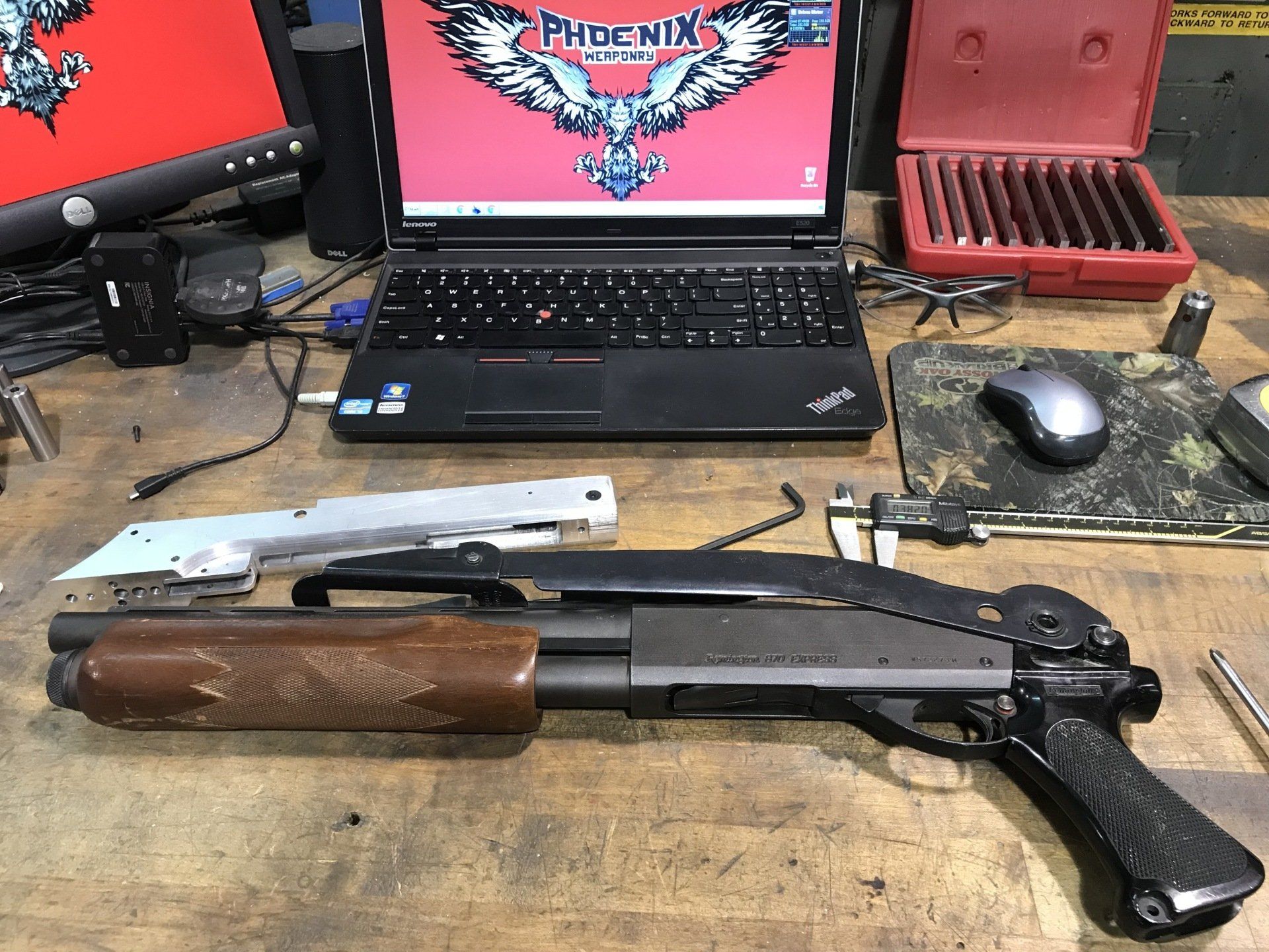 Shotgun with a wooden grip on a workbench, computer and tools in the background.