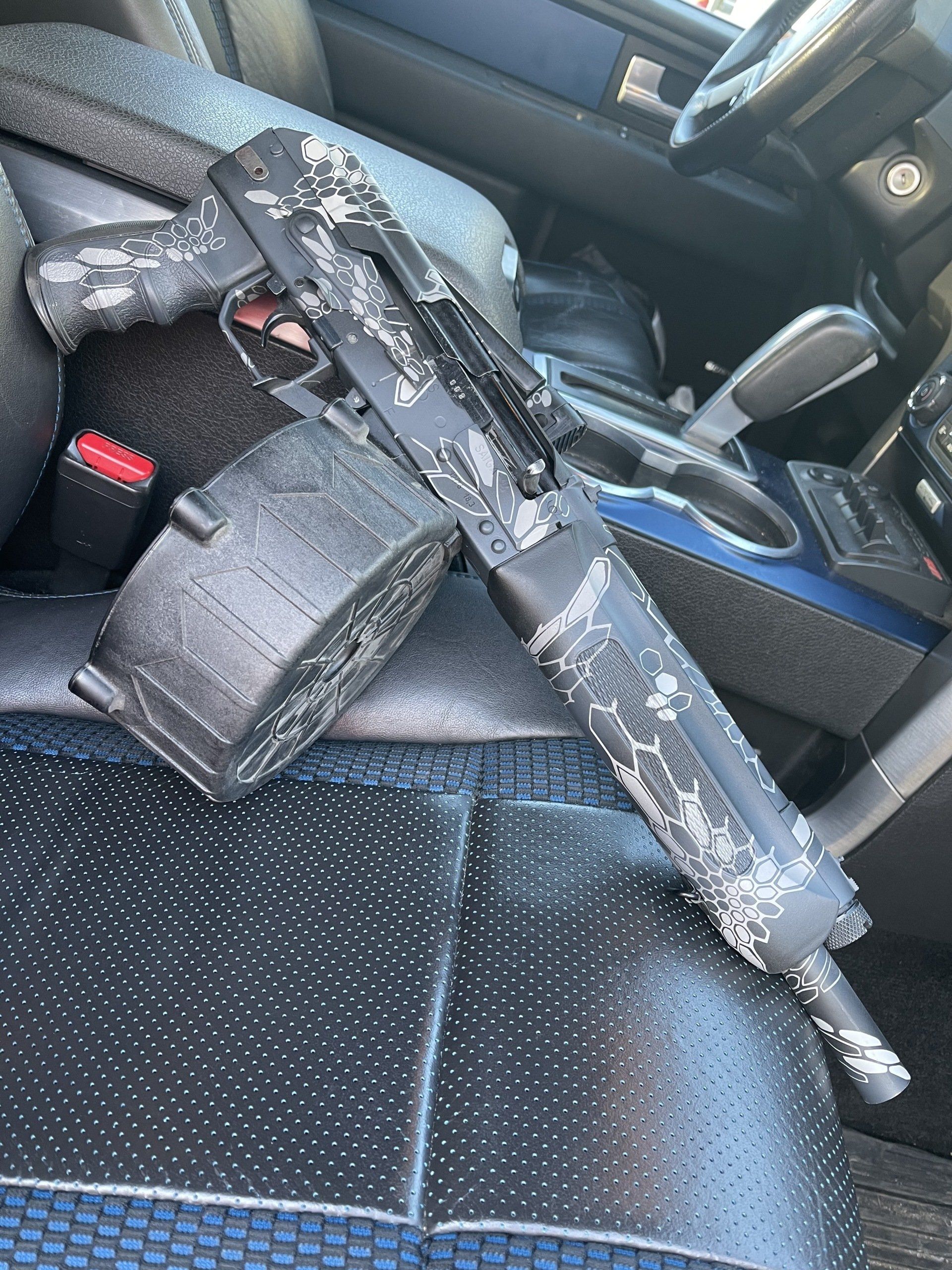 Black firearm with drum magazine on vehicle seat.