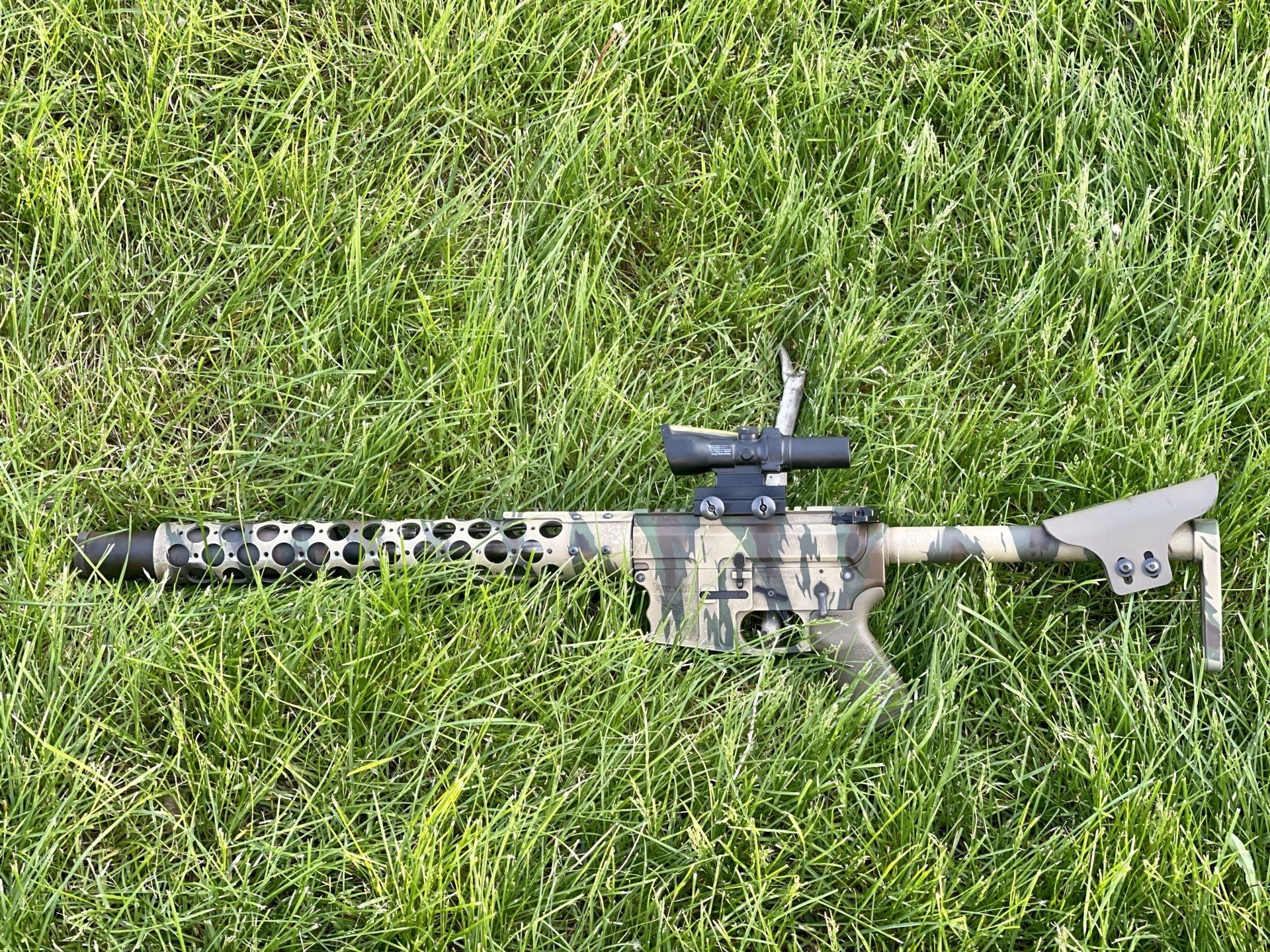 Camouflaged rifle resting in green grass, equipped with a scope.