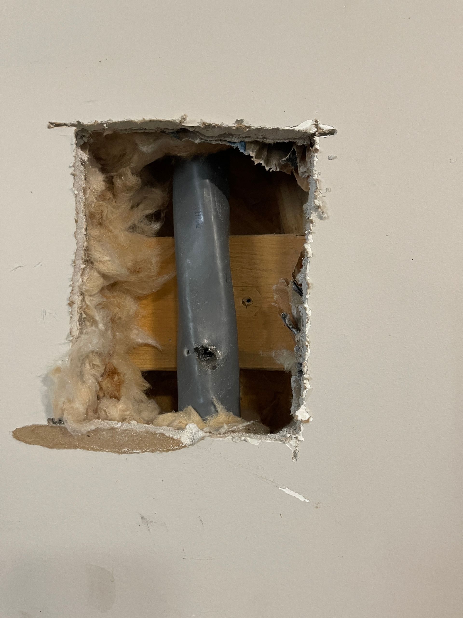 Hole in wall exposing insulation and a vertical gray pipe. Wood support beams visible behind pipe.