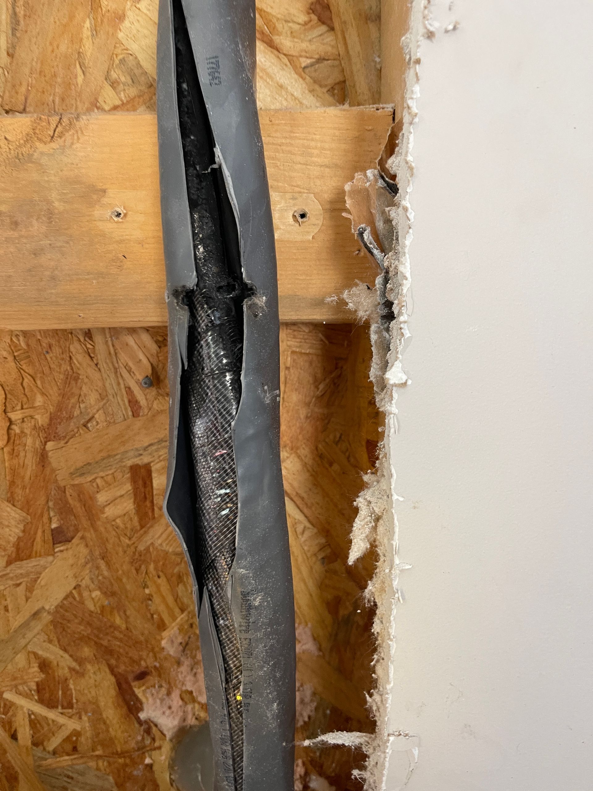 Damaged black electrical cable exposed between wooden panel and white wall, showing inner wires.