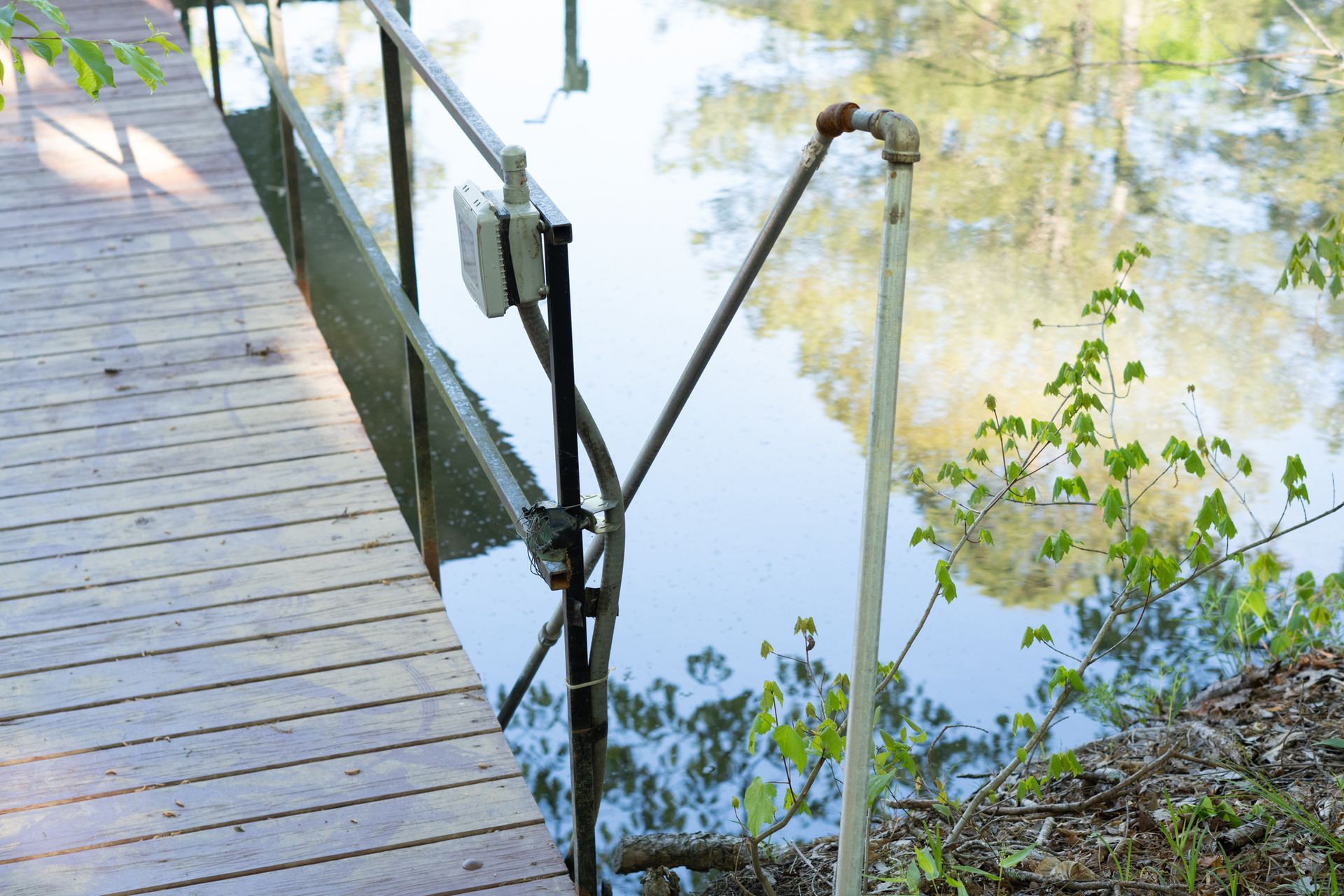 Wooden dock extending into water with metal railing and pipe. Green foliage and water visible.