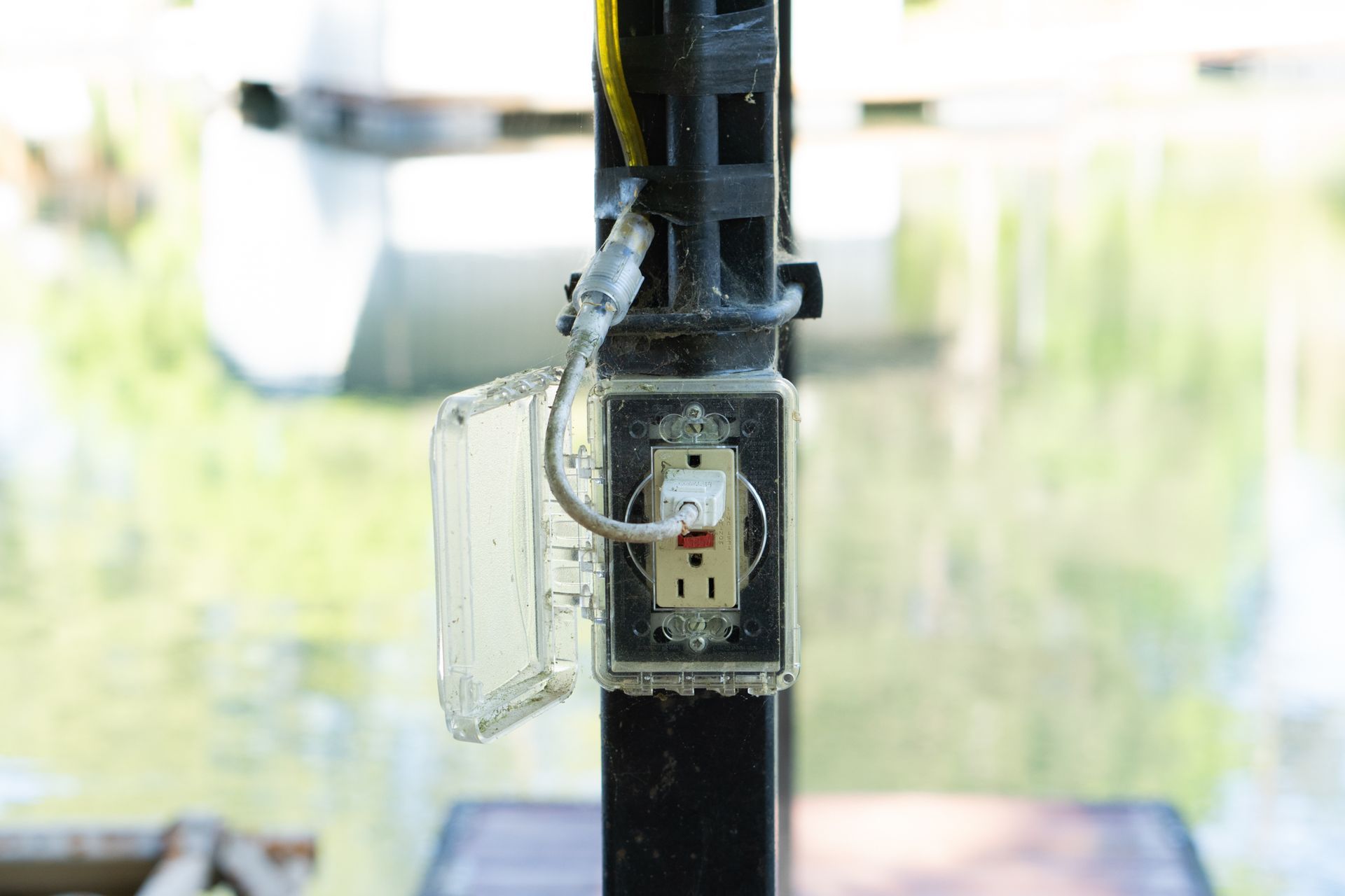 Outdoor electrical outlet with protective cover, mounted on a black post, with water in the background.