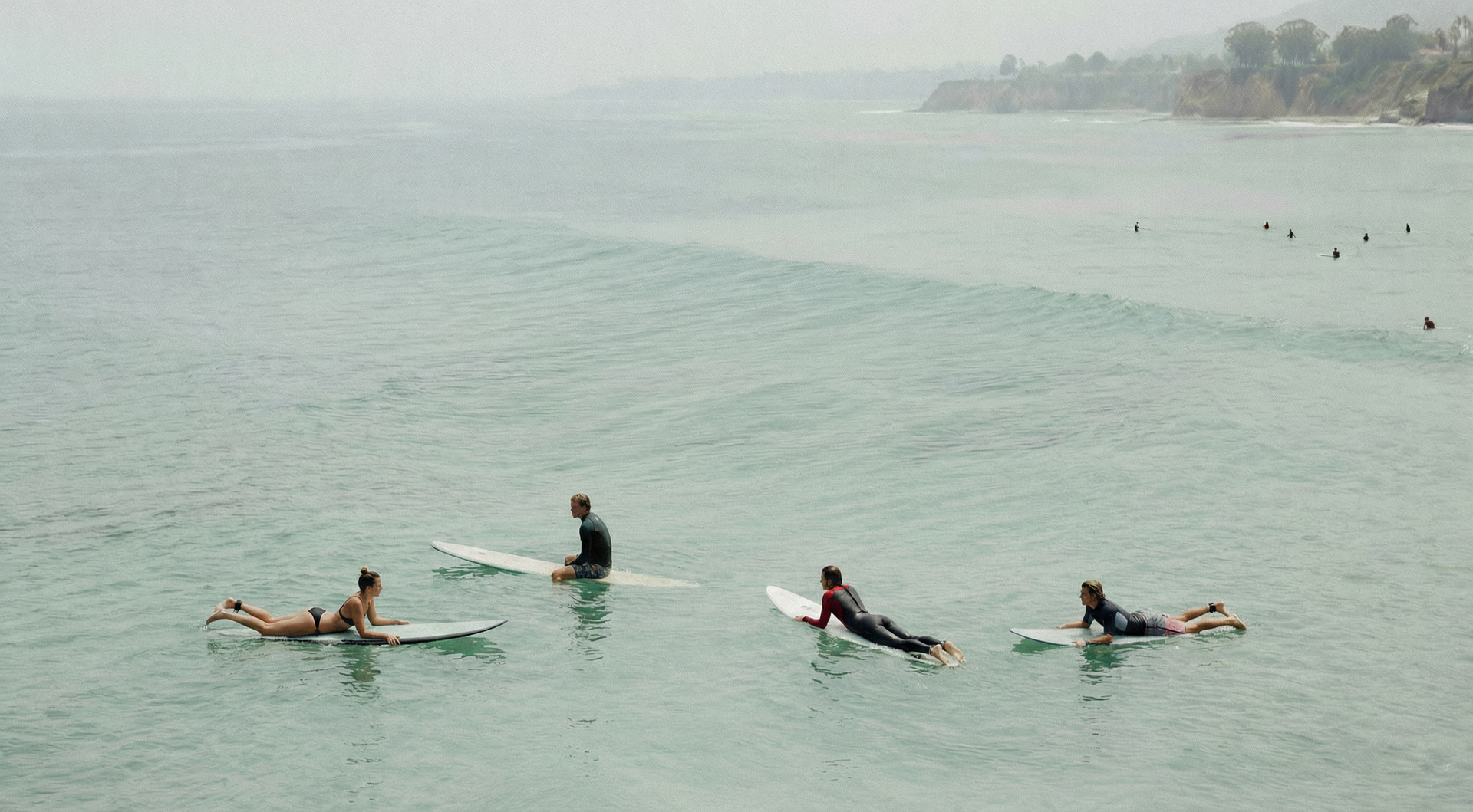 An aerial view of two surfers on turquoise water, one waiting on a board while the other carves a turn on a wave.