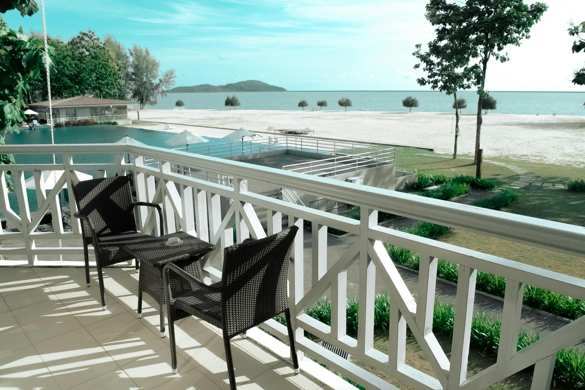 A balcony with two dark wicker chairs overlooking a swimming pool, sandy beach, and the sea on a sunny day.