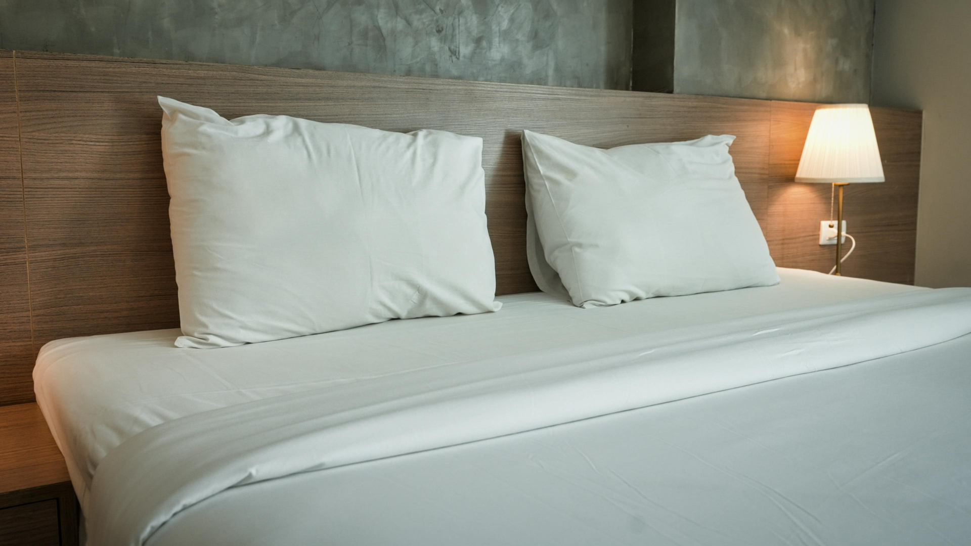 Neatly made bed with white pillows and lamp beside a wooden headboard in a hotel room