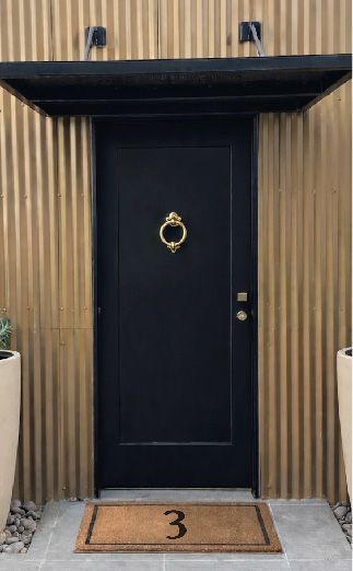 A black front door with a gold knocker, positioned beneath a black awning against a vertical, corrugated metal wall.