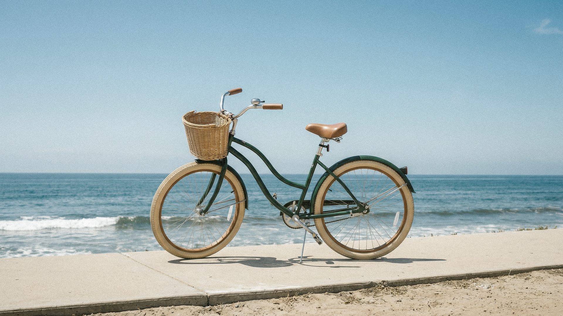 A person smiling while riding a white electric bicycle on a paved beach path with sand and palm trees in the background.