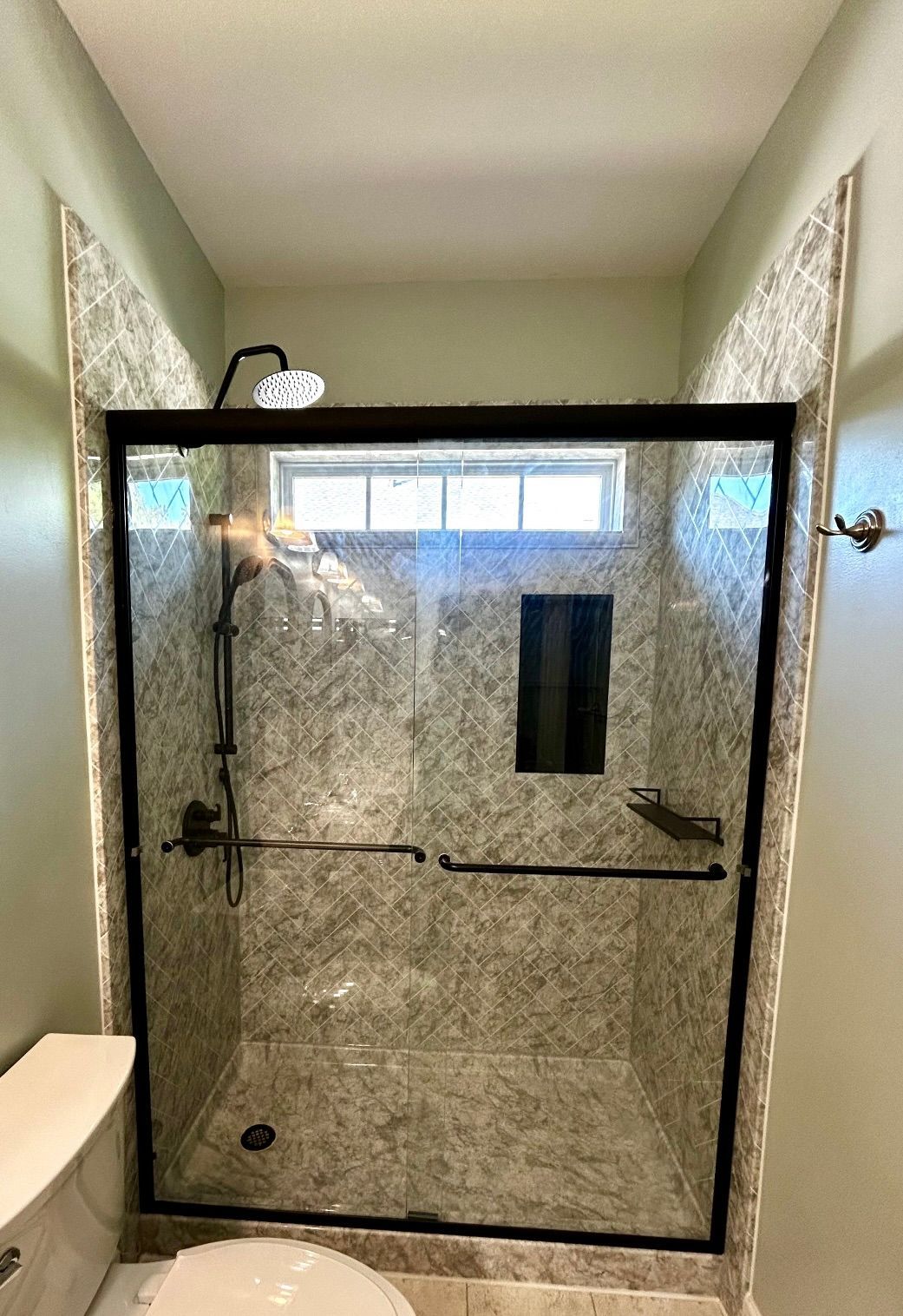 Glass shower enclosure in a narrow tiled bathroom with a window above.