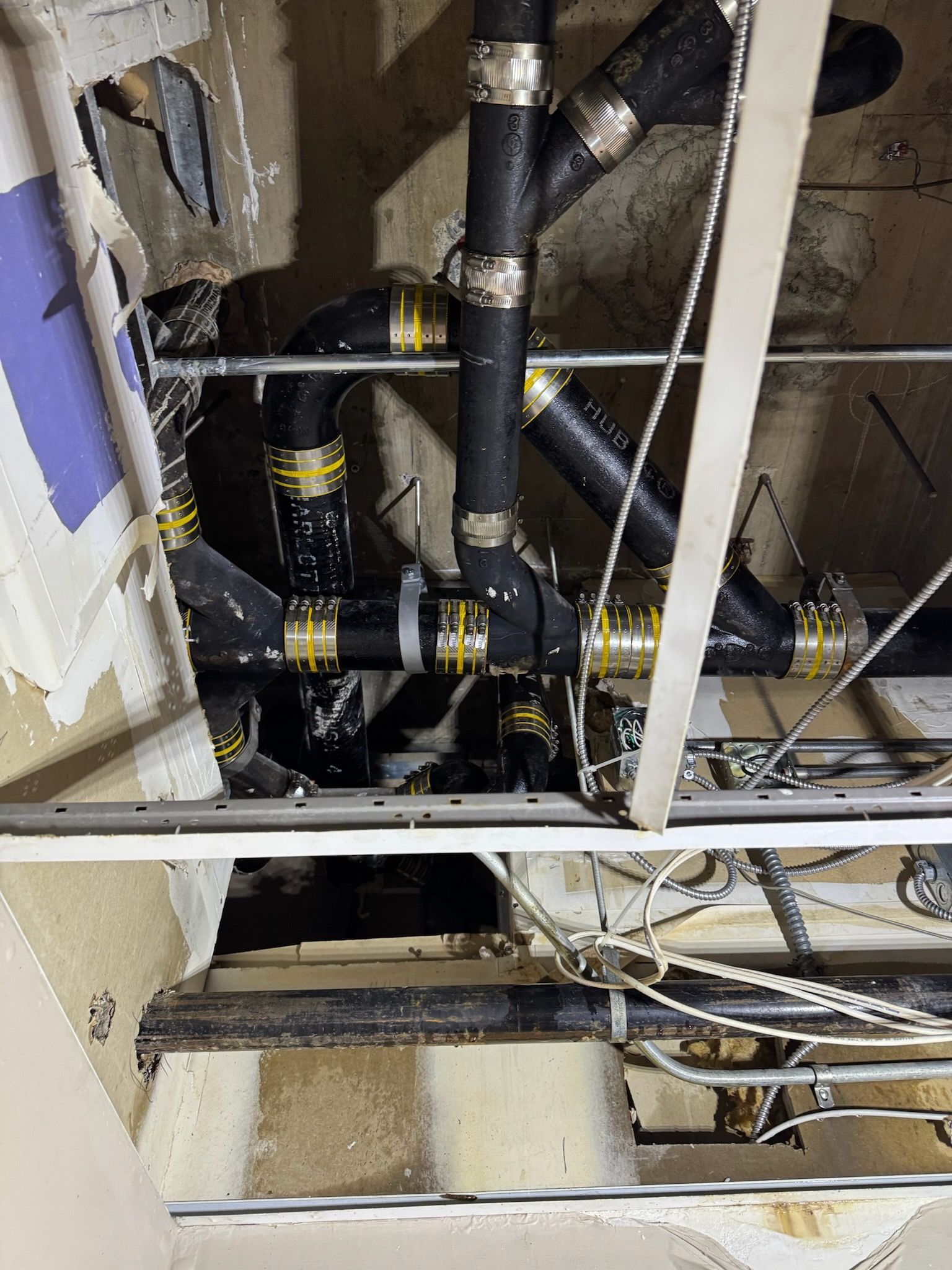 Exposed black plumbing pipes and white beams inside a ceiling crawlspace, with cables and insulation.
