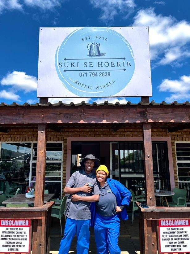 Two people smiling in front of a restaurant sign, 