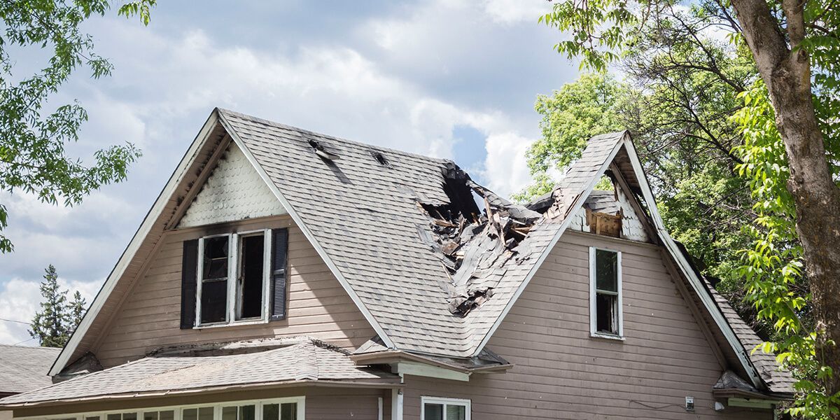 House with extensive roof damage, likely from a fire. Burnt roof, broken windows, and debris are visible.