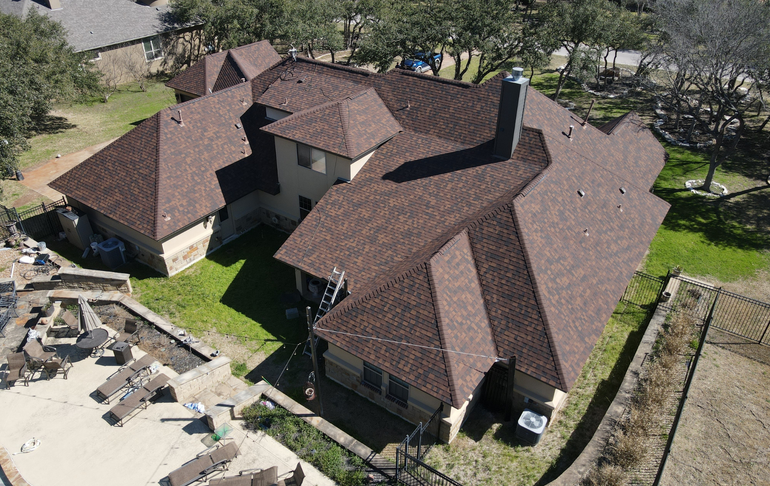 Aerial view of a brown-roofed house with multiple sections, surrounded by green lawn and trees.