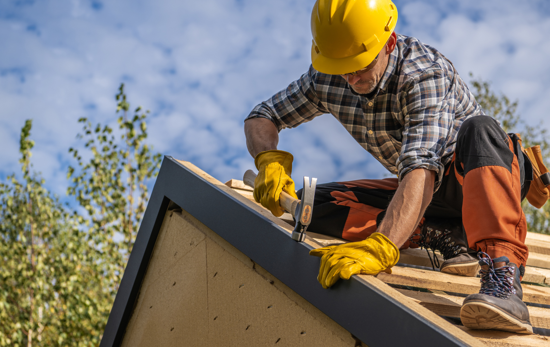 Roofer in yellow hard hat hammers on a roof, wearing gloves and orange overalls, outdoors on a sunny day.