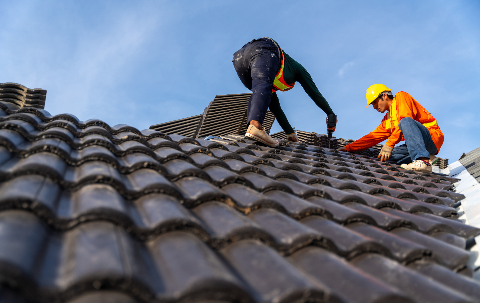 Roofer on a roof, securing shingles with a tool, wearing a safety harness and tool belt.