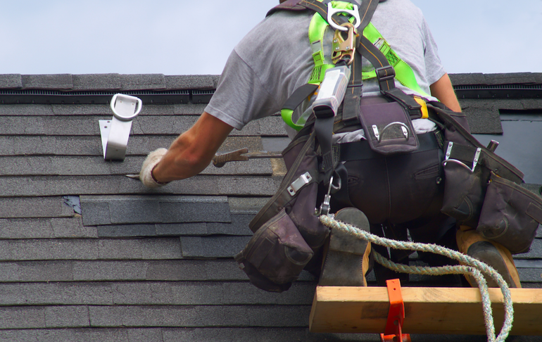 Roofer on a roof, securing shingles with a tool, wearing a safety harness and tool belt.