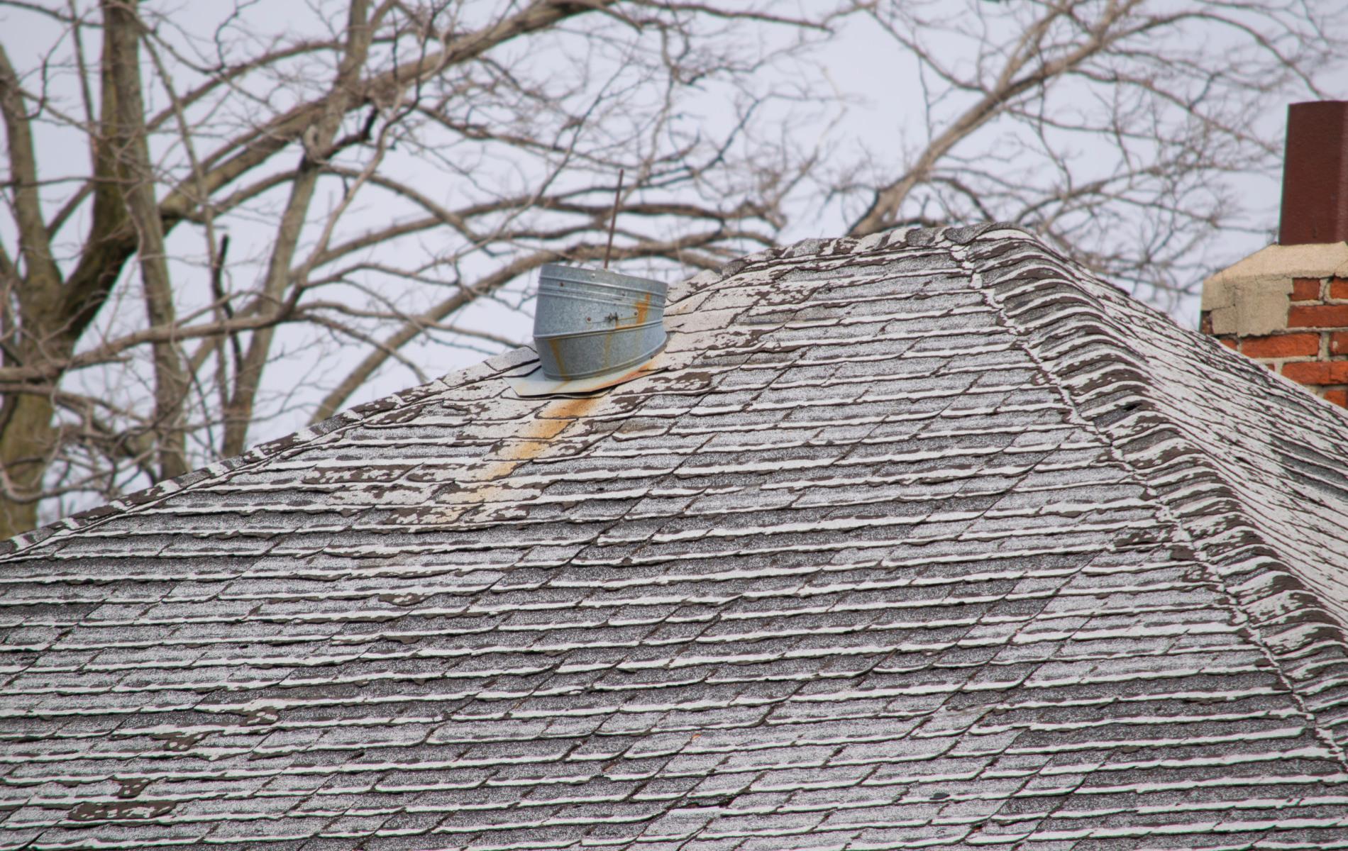 Weathered roof with a metal vent and brick chimney against leafless trees.