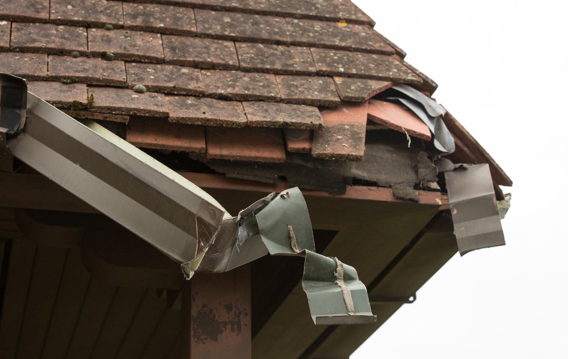 Damaged roof with missing tiles, crumpled gutter, against a cloudy sky.