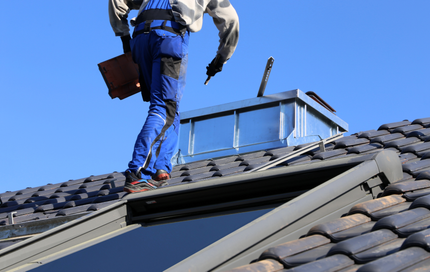 Roofer in blue overalls on a tiled roof near a skylight, with a blue sky background.