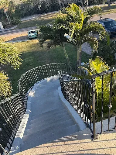 Curved outdoor staircase with black railings descending past palm trees toward a street and parked cars