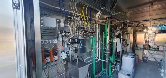 Attrezzatura industriale con pompe, manometri e tubi; uno sfondo con pannello metallico.
