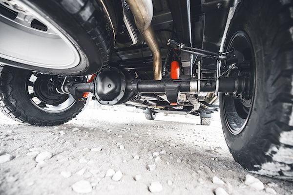 Underbody view of a vehicle’s suspension and axles on snowy ground, with a visible tire and orange component
