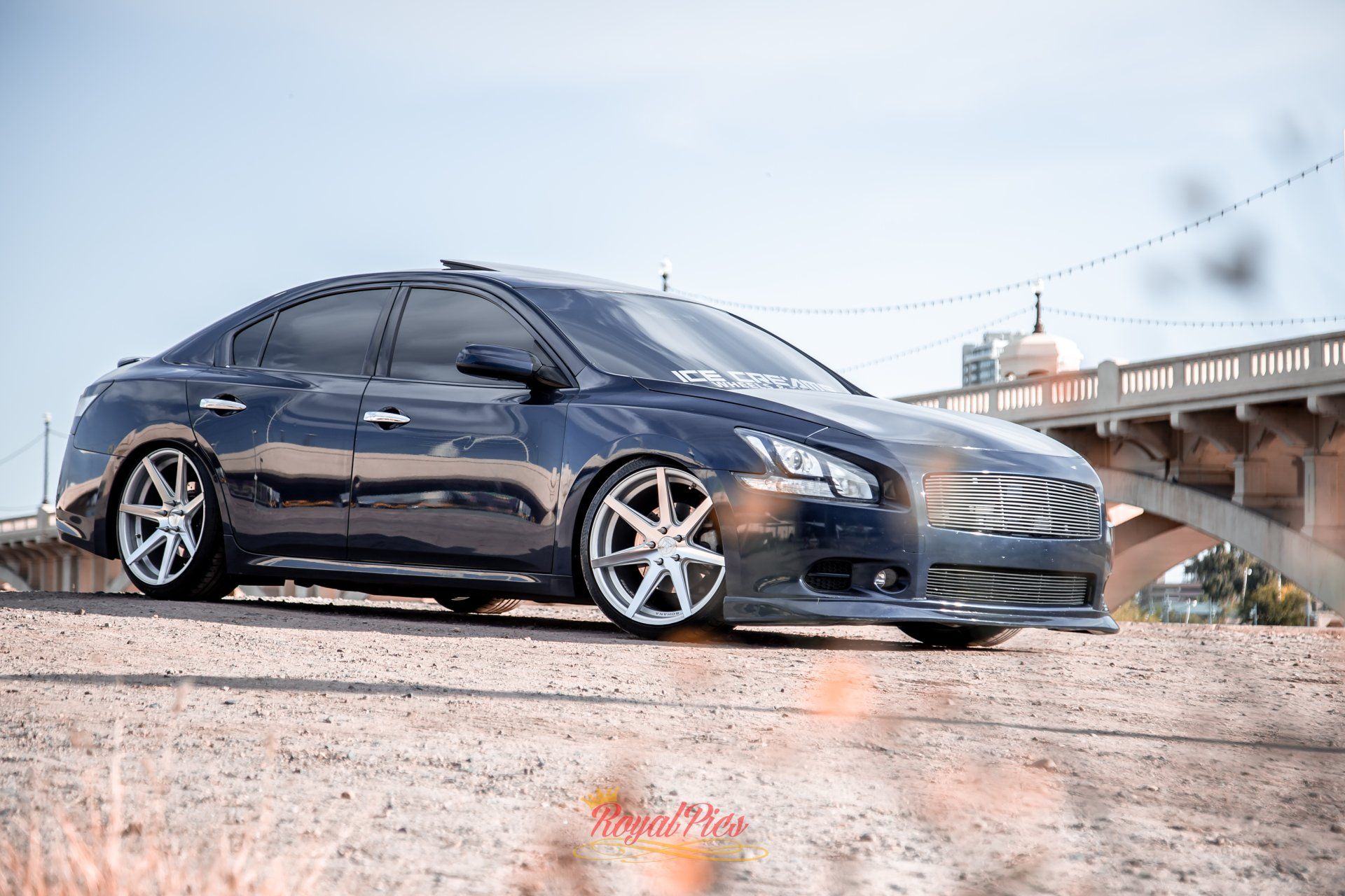Glossy black sedan with chrome rims parked outdoors near a bridge at sunset