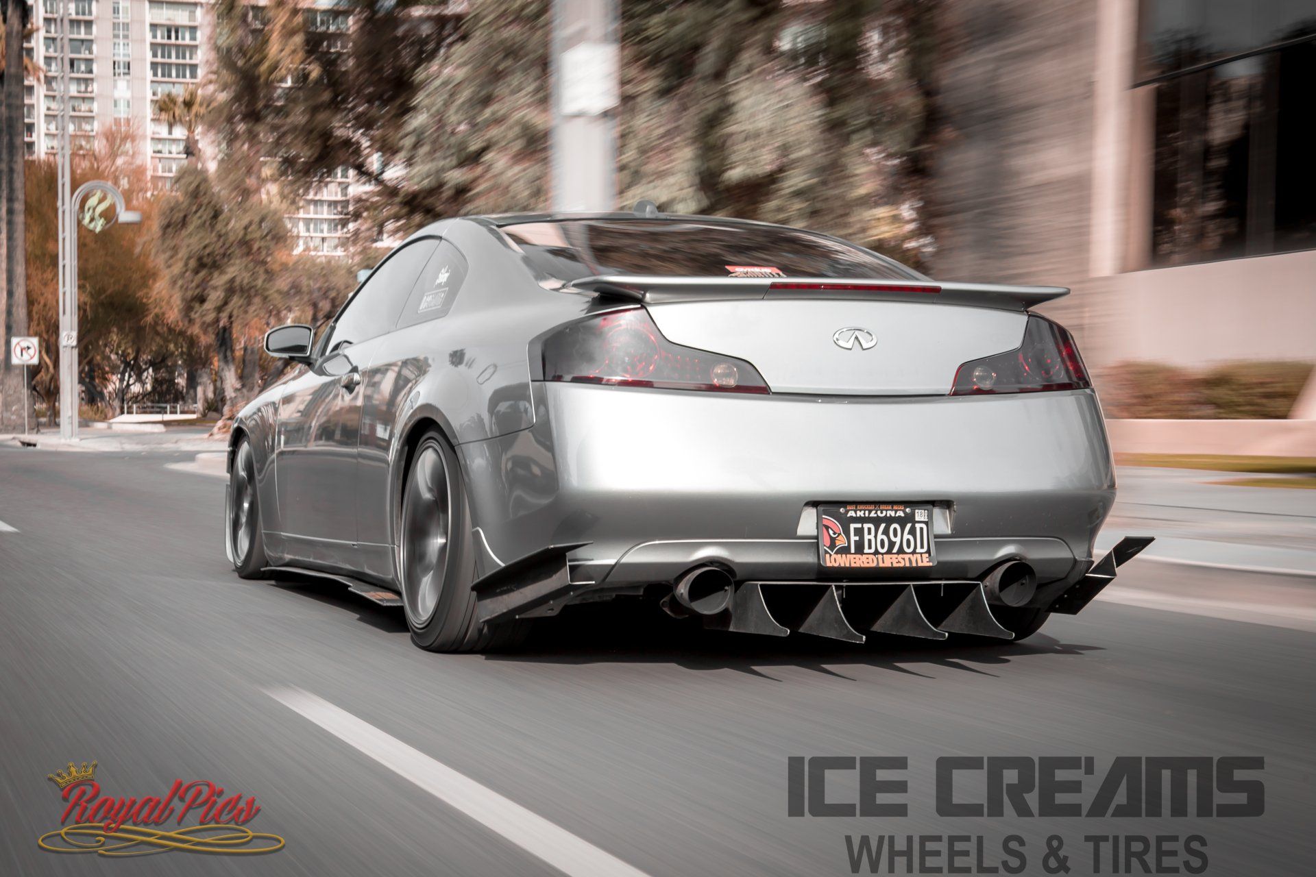 Silver coupe driving on a city street with wide rear wheels and an aftermarket body kit