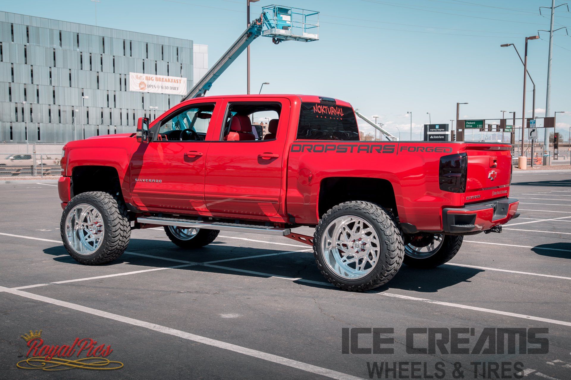 Red lifted pickup truck in a parking lot, viewed from the side with custom wheels.