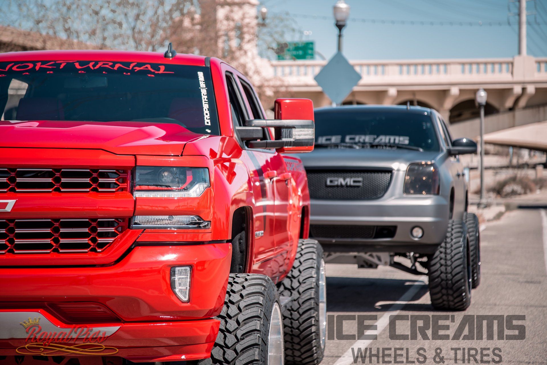 Red lifted pickup truck in front of a gray GMC truck on a city street with overpass and tire shop sign