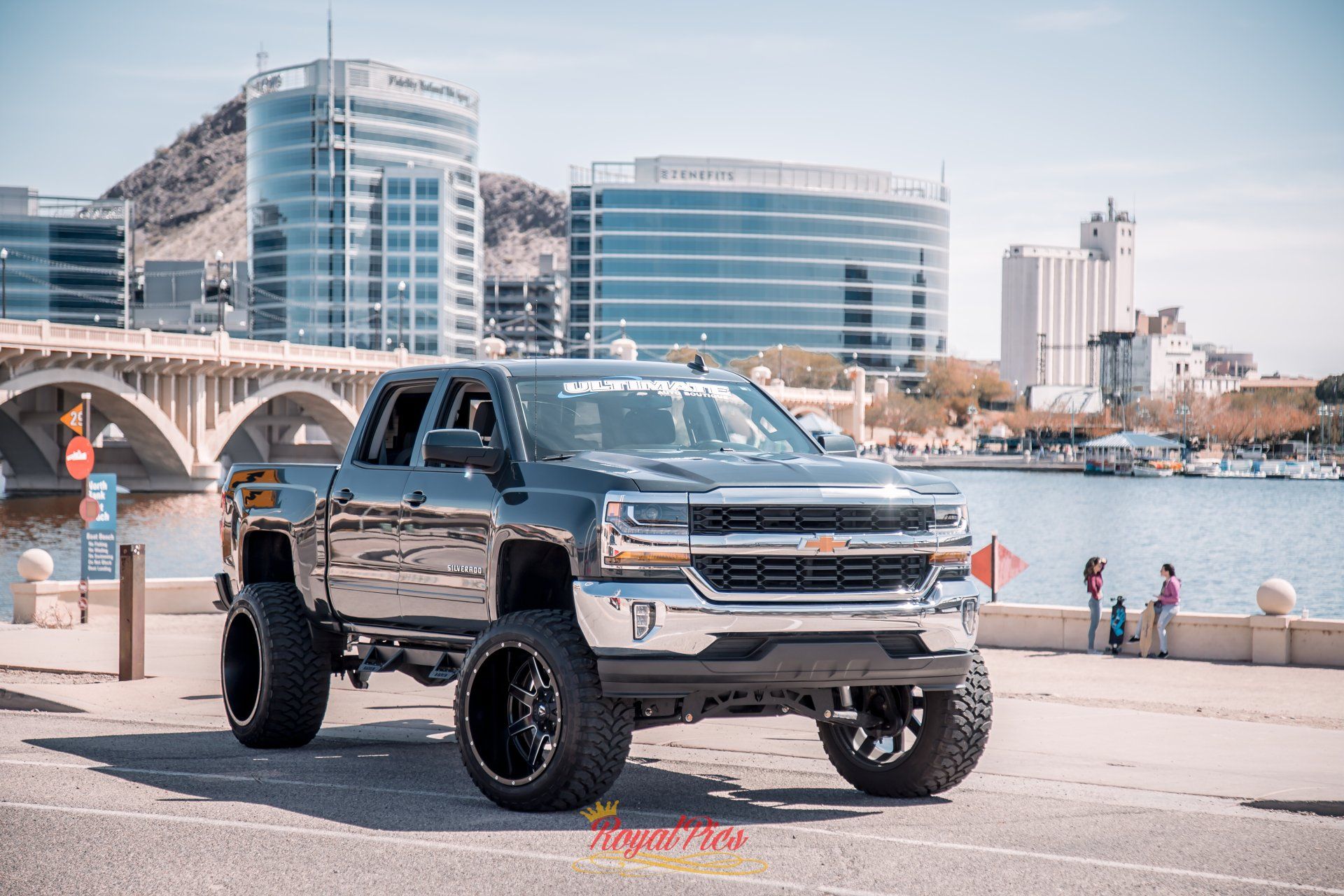 Lifted black pickup truck parked by a waterfront bridge with modern city buildings in the background