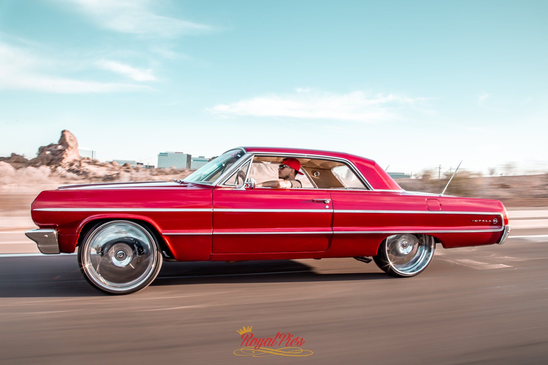 Red classic car driving on a road under a bright blue sky.