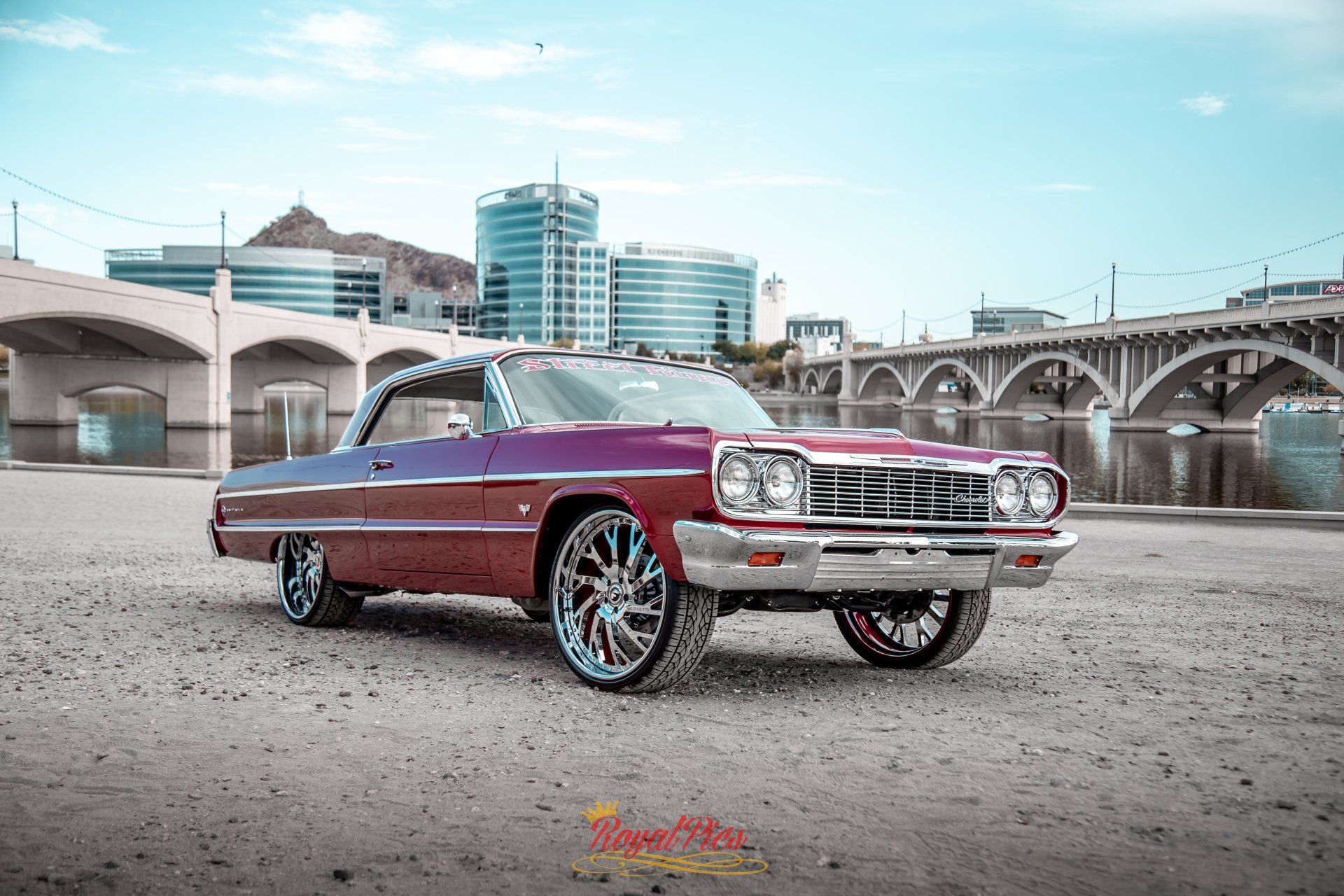 Classic burgundy lowrider car parked by a riverfront bridge and city skyline