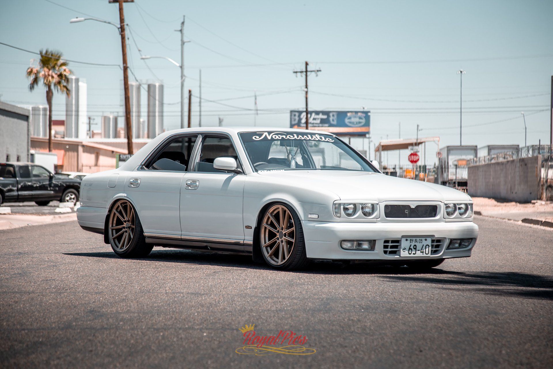 White lowered sedan parked on a street, with bronze wheels and a sunny urban background