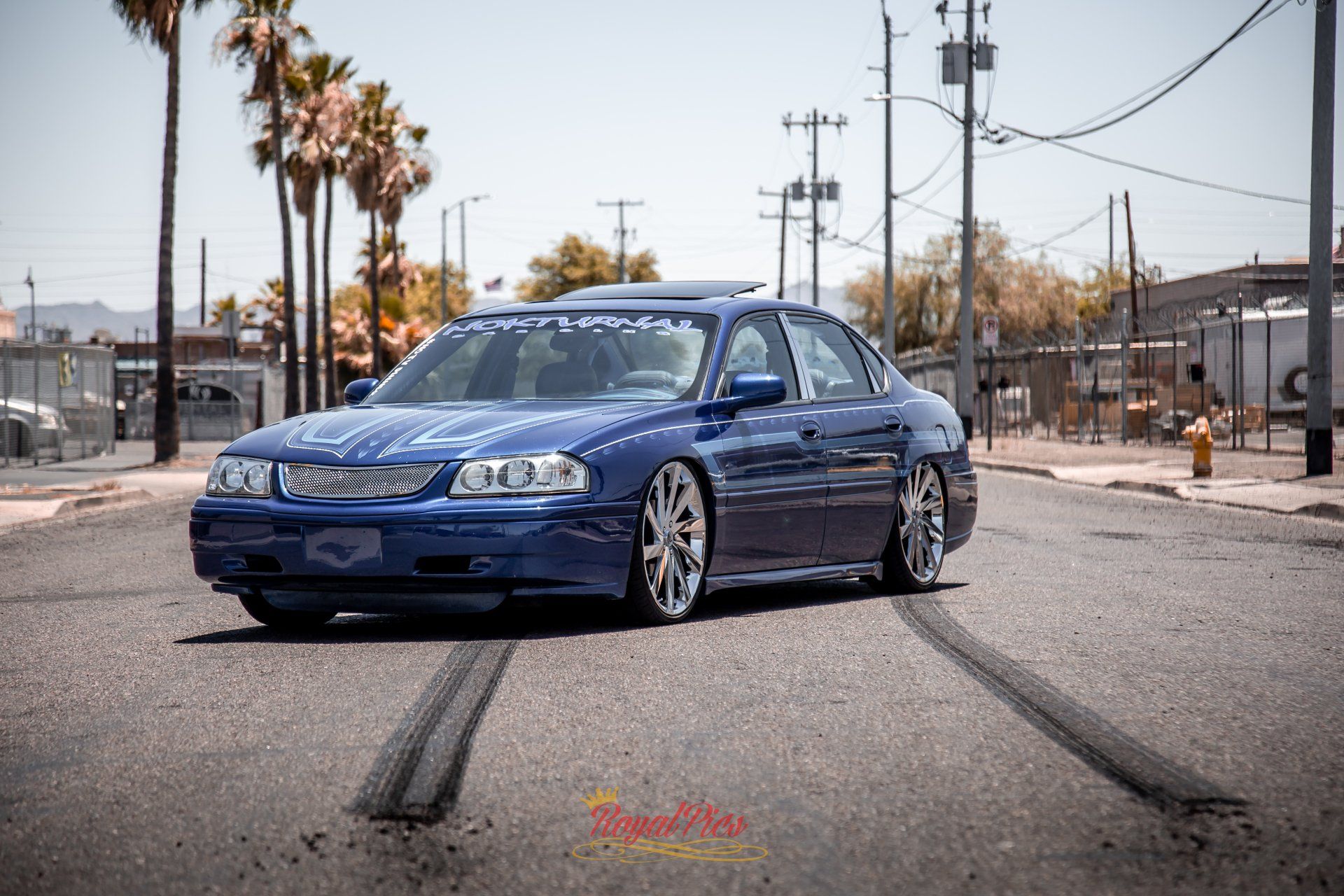 Blue sedan parked on a street with dark tire marks, palm trees and utility poles in the background