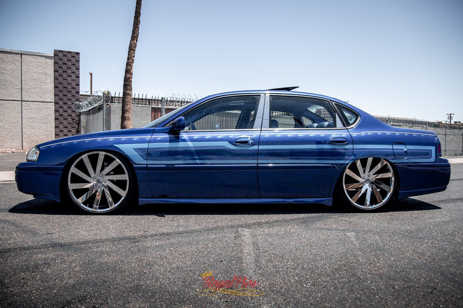 Blue lowered sedan parked on a street in front of a brick building and chain-link fence.