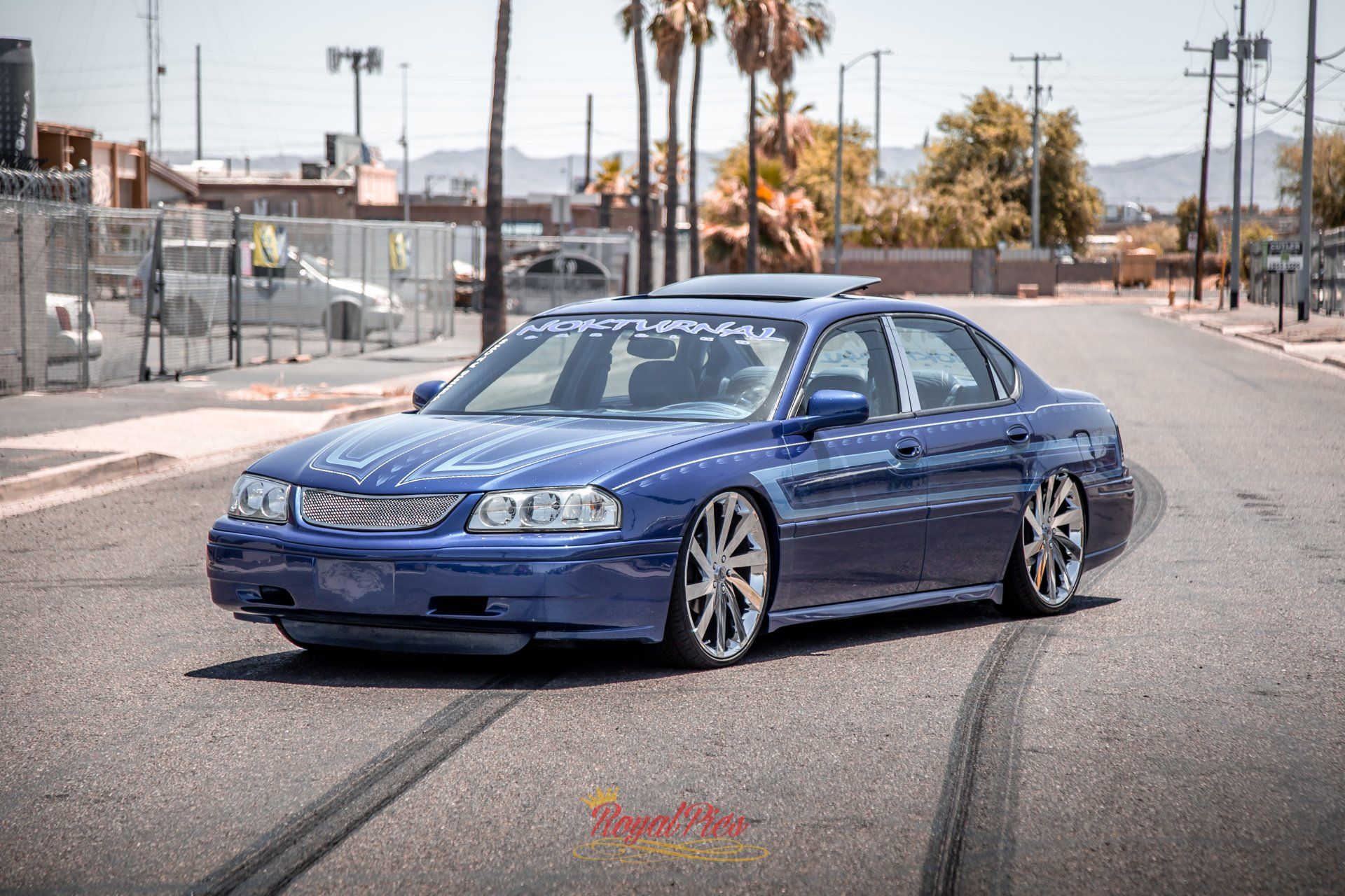 Blue sedan parked on an empty street near a chain-link fence and palm trees under clear daylight