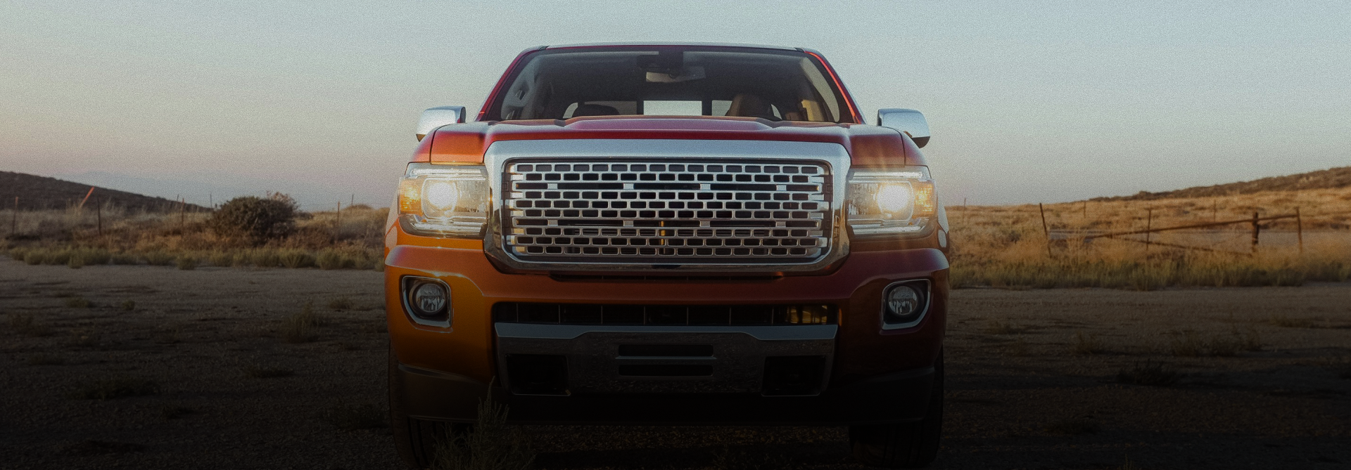 Orange pickup truck facing forward on a road at sunset, with headlights on and open landscape behind it