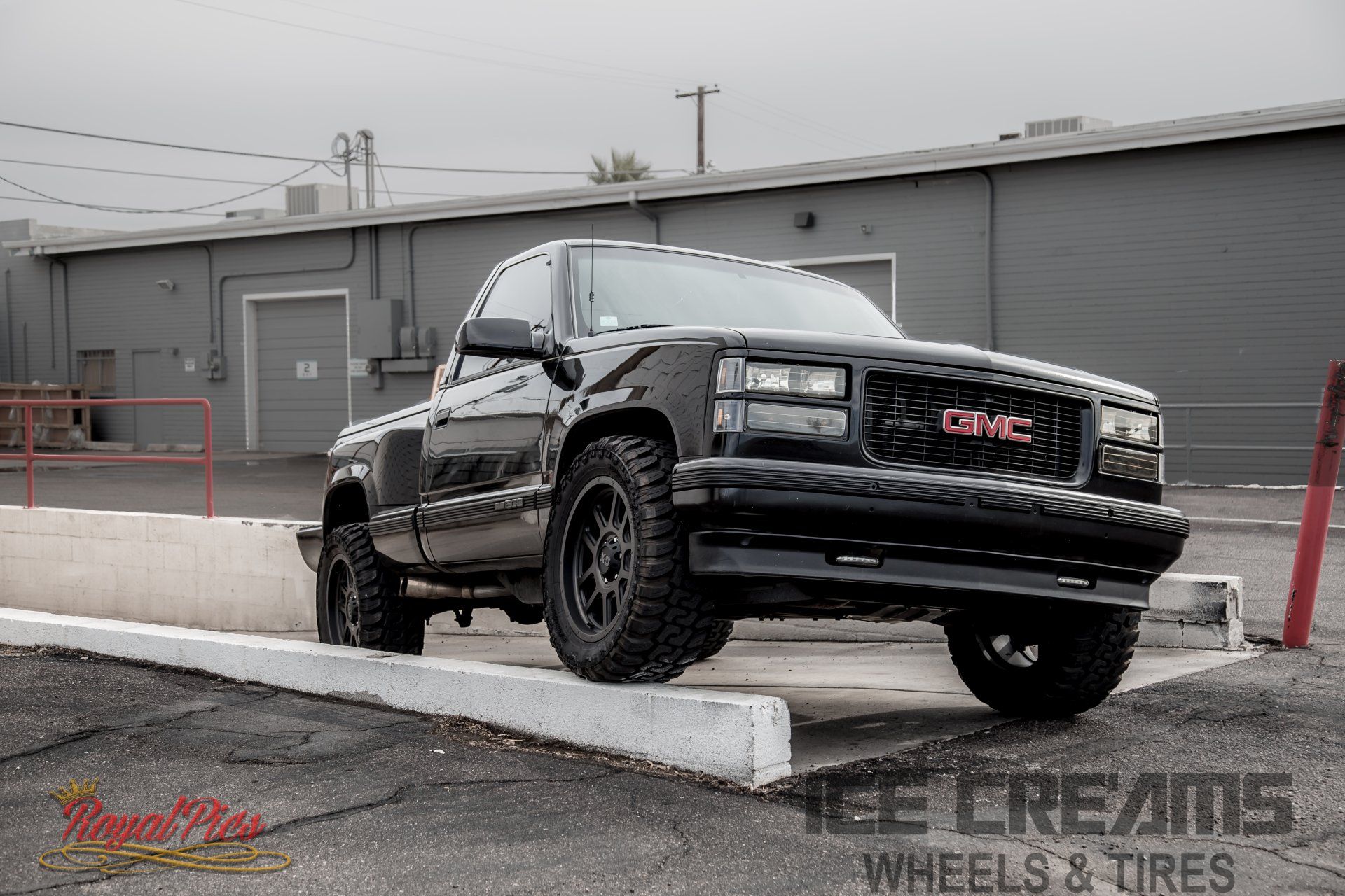 Black GMC pickup truck parked on a concrete curb in front of a gray industrial building