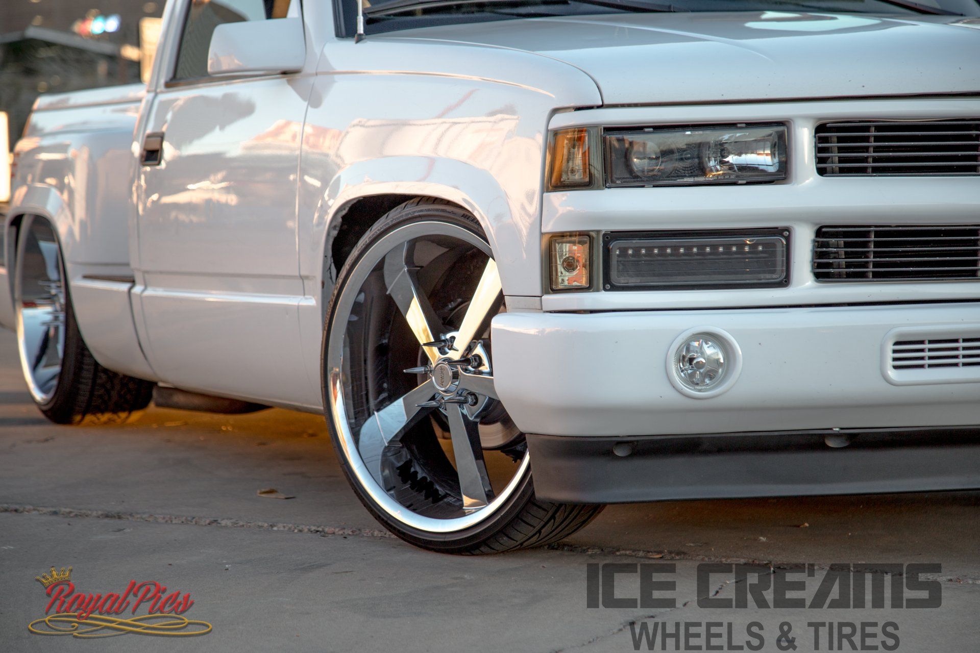 Silver custom pickup truck with chrome rims parked in front of a shop, close-up on the front wheel.