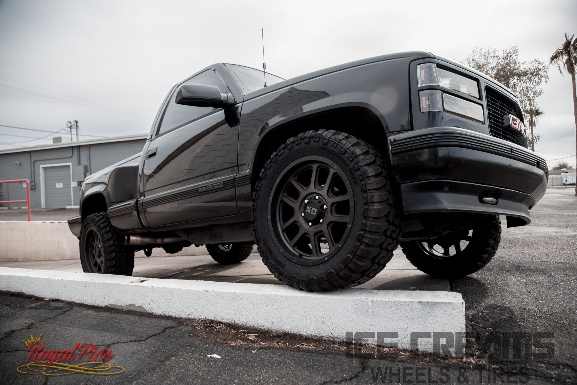 Lifted black pickup truck with off-road tires parked on a curb in an industrial lot