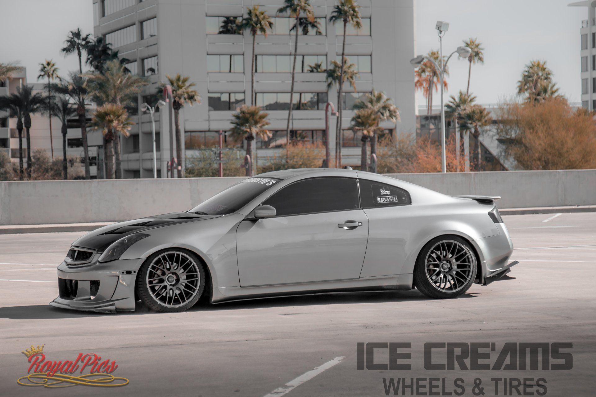 Gray lowered coupe with black wheels parked in a lot, palm trees and apartment building in the background